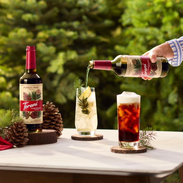 Lifestyle image of Torani Puremade Forest Pine Syrup being poured into a pine-garnished drink outdoors